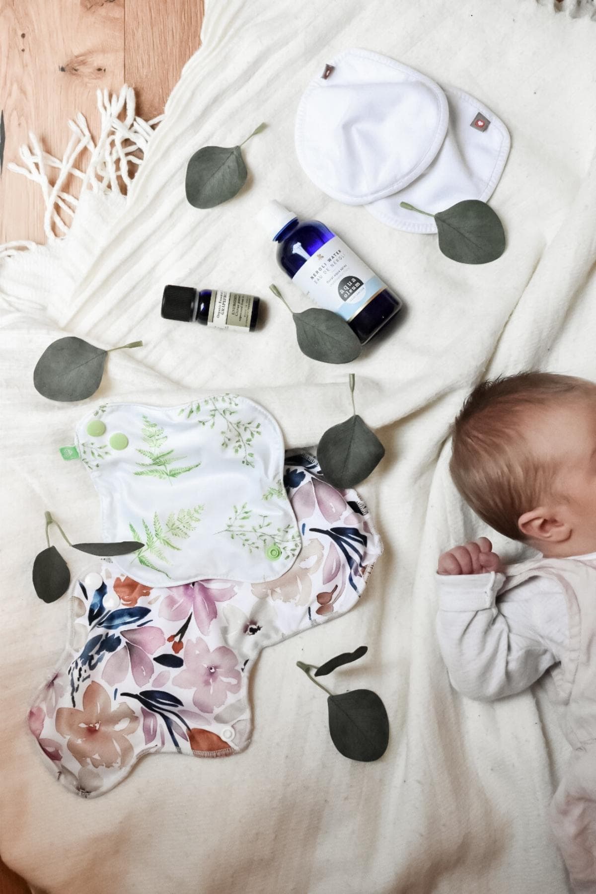 A baby lies on a white blanket surrounded by greenery, epitomizing a comfortable birth recovery. Nearby are reusable floral-printed and leaf-patterned pads, two bottles of skincare products, and two round white cloth pads. Scattered eucalyptus leaves complete the tranquil scene.