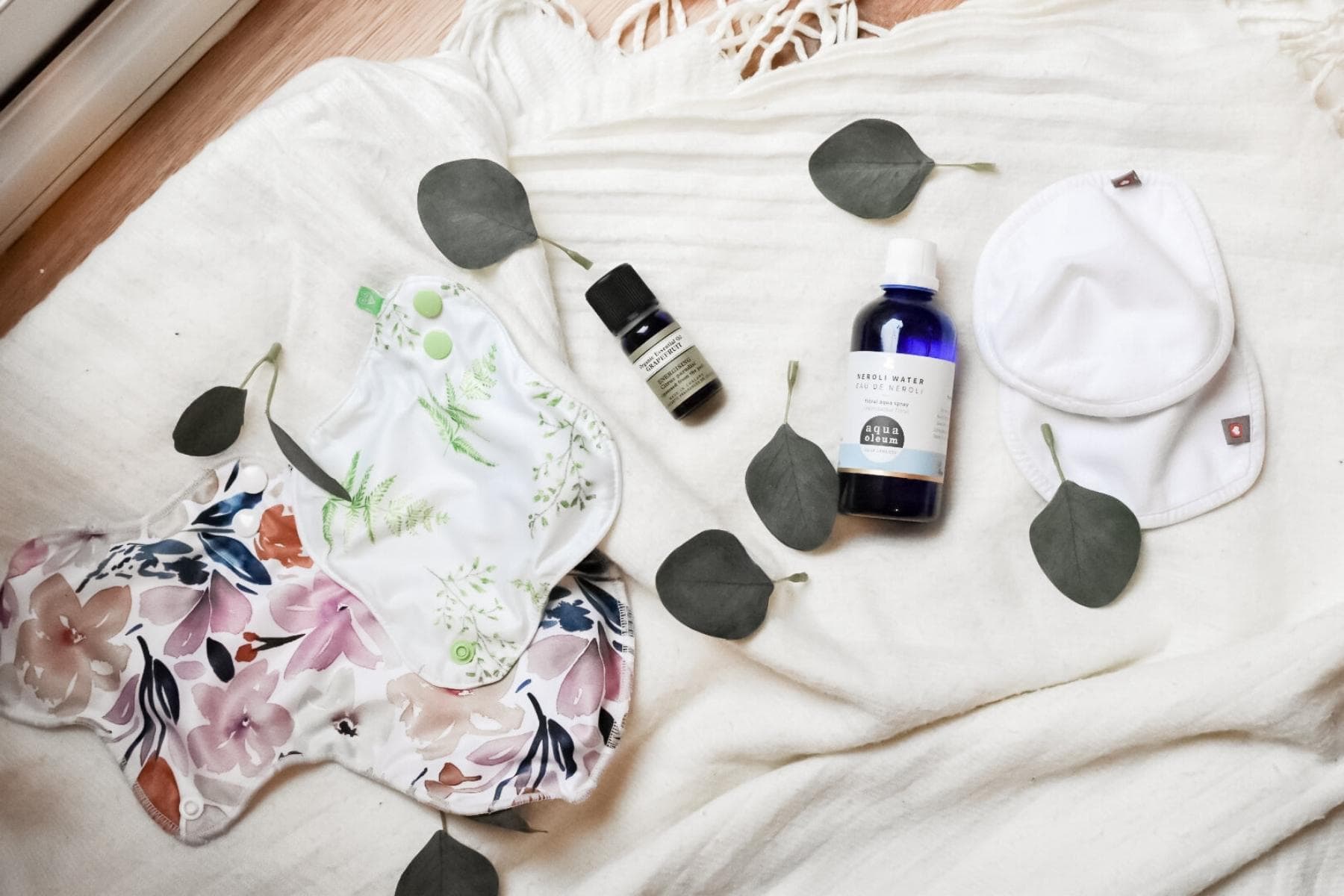 Cloth sanitary pads featuring floral and leaf patterns are spread out on a white blanket. Perfect for sustainable birth recovery, they are accompanied by a small essential oil bottle and a larger witch hazel bottle, scattered among green leaves.