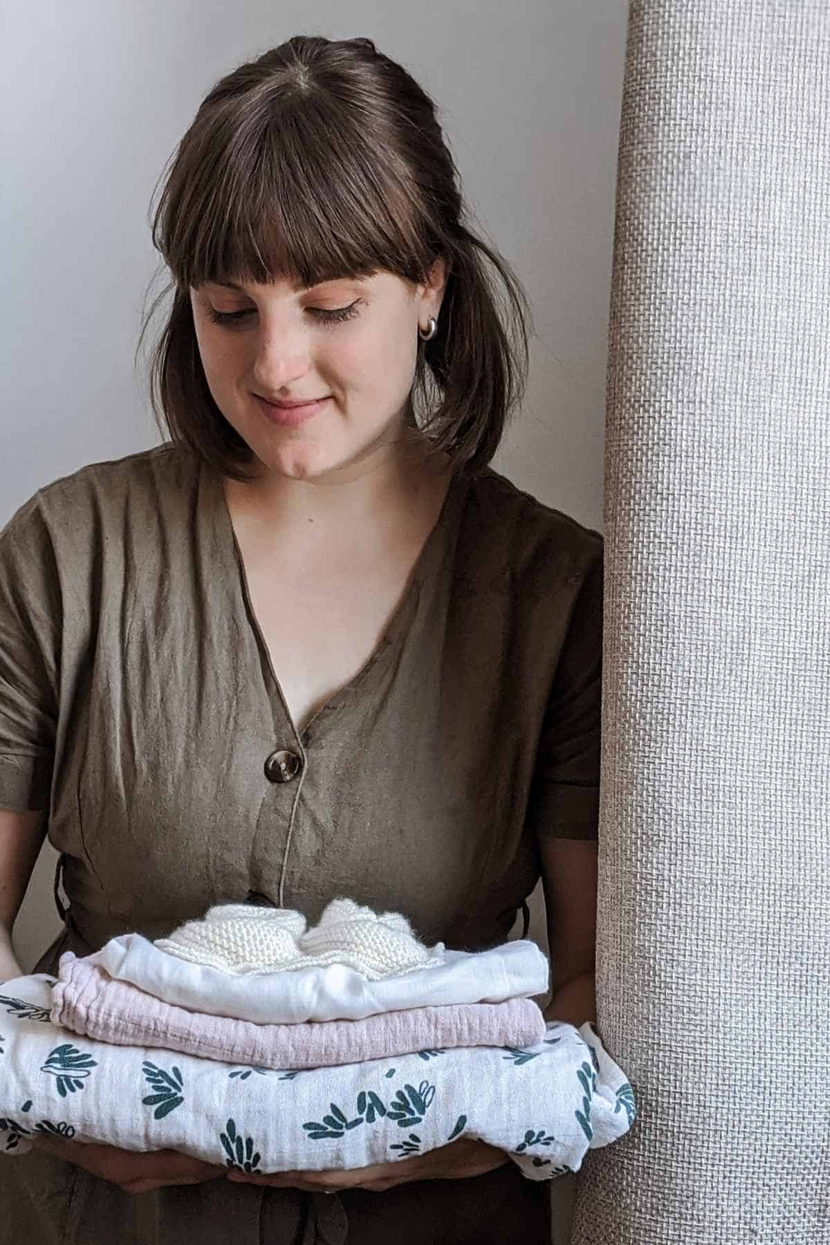 A person with shoulder-length brown hair, wearing a sustainable dress, stands near a beige curtain. They hold a folded stack of ethical breastfeeding clothes, including a white sweater and patterned fabric with green leaves.