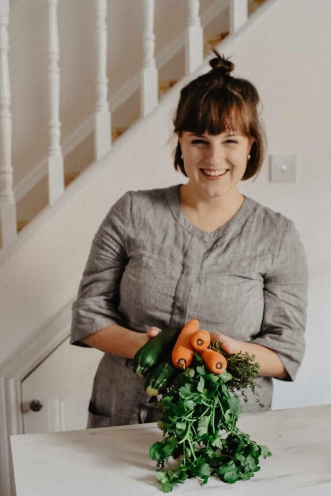 A person with short brown hair stands indoors, smiling and holding a bunch of fresh vegetables, including carrots, zucchini, and leafy greens, embracing a sustainable lifestyle in front of a white staircase railing. They are wearing a gray top.