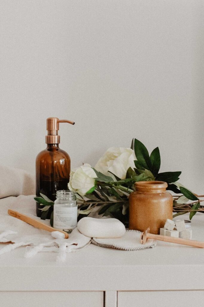 Aesthetic arrangement of eco-friendly skincare products on a white surface. Includes a brown glass pump bottle, a small jar, a tan jar, leaves, and white flowers. Brushes and a soap dish complement the natural, serene setting—subtle nods to a sustainable lifestyle with small changes in packaging choices.