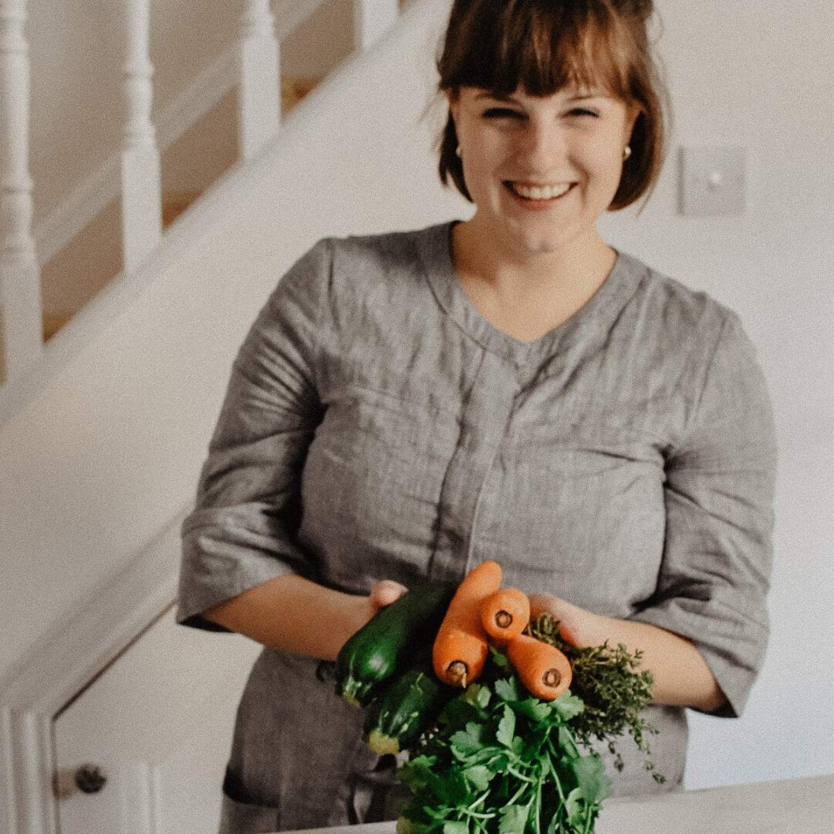 A person in a gray outfit stands indoors, smiling and embracing a sustainable lifestyle by holding fresh vegetables like carrots and zucchini, along with herbs. Small changes in daily habits can make a big difference for the planet.