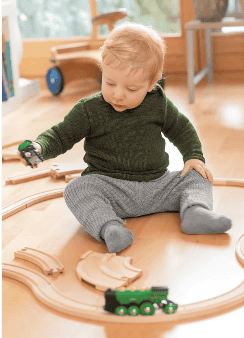 A toddler with blond hair, wearing sustainable children's clothes - a green sweater and gray pants by Disana. He sits on a wooden floor playing with a wooden train set. The child is holding a piece of the train, surrounded by tracks and toys in a cozy, light-filled room.