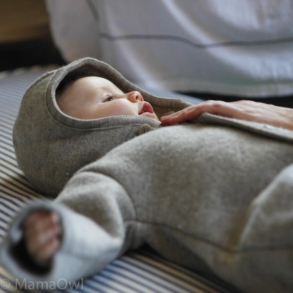 A baby wearing a gray hooded Disana onesie lies on a striped surface. A hand gently rests on the chest of the baby, who is looking upwards with a calm expression.