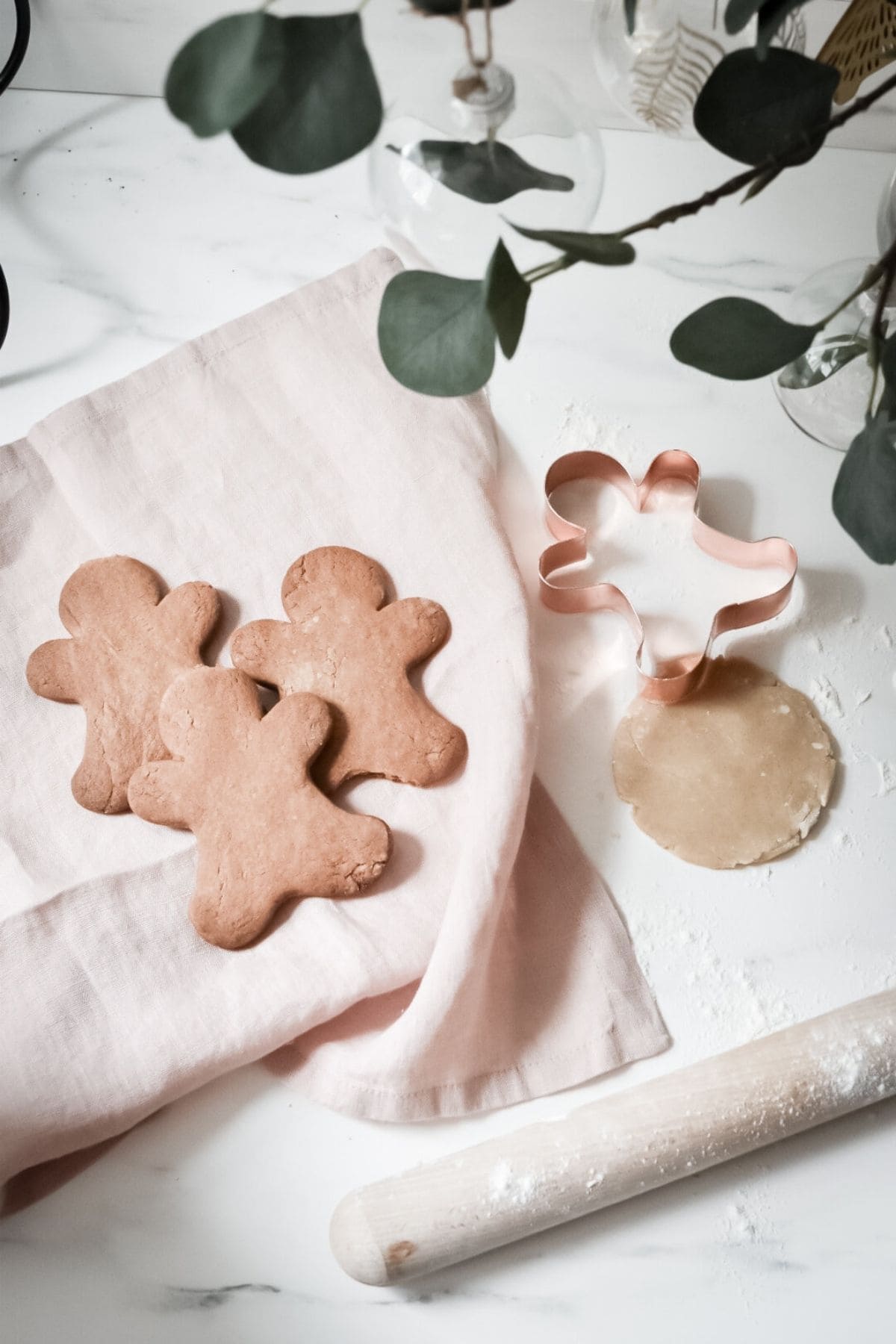 Three gingerbread cookies shaped like people rest on a pink cloth. Nearby, a gingerbread-shaped cutter sits on rolled-out dough. A rolling pin and eucalyptus leaves enhance the scene, embodying the spirit of an eco-friendly holiday on a crisp white surface.