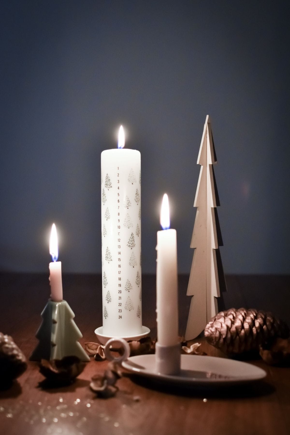 A cozy scene unfolds with three lit candles of varying heights and shapes on a wooden surface. Decorative pinecones and a wooden tree figurine add an eco-friendly holiday touch.