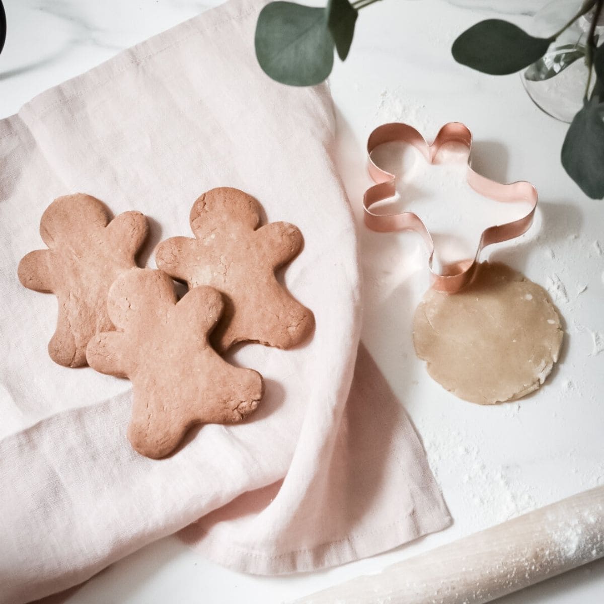 Three gingerbread cookies rest on a light pink cloth. Nearby, a copper gingerbread man-shaped cookie cutter sits next to rolled dough on a white surface, surrounded by green leaves—creating the perfect sustainable Christmas scene.