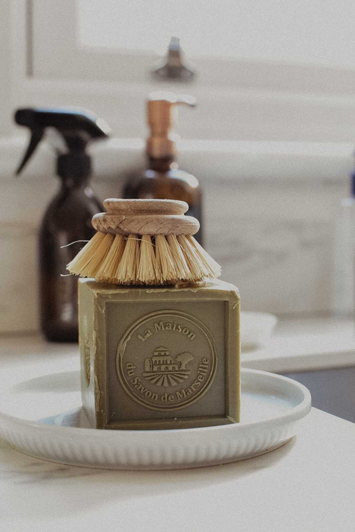 A block of green soap with Le Maison du Savon de Marseille engraved on it sits on a white dish, symbolizing a sustainable lifestyle. A round scrub brush rests on top. In the background, spray and pump bottles adorn the countertop near a window, reflecting post-pandemic cleanliness.
