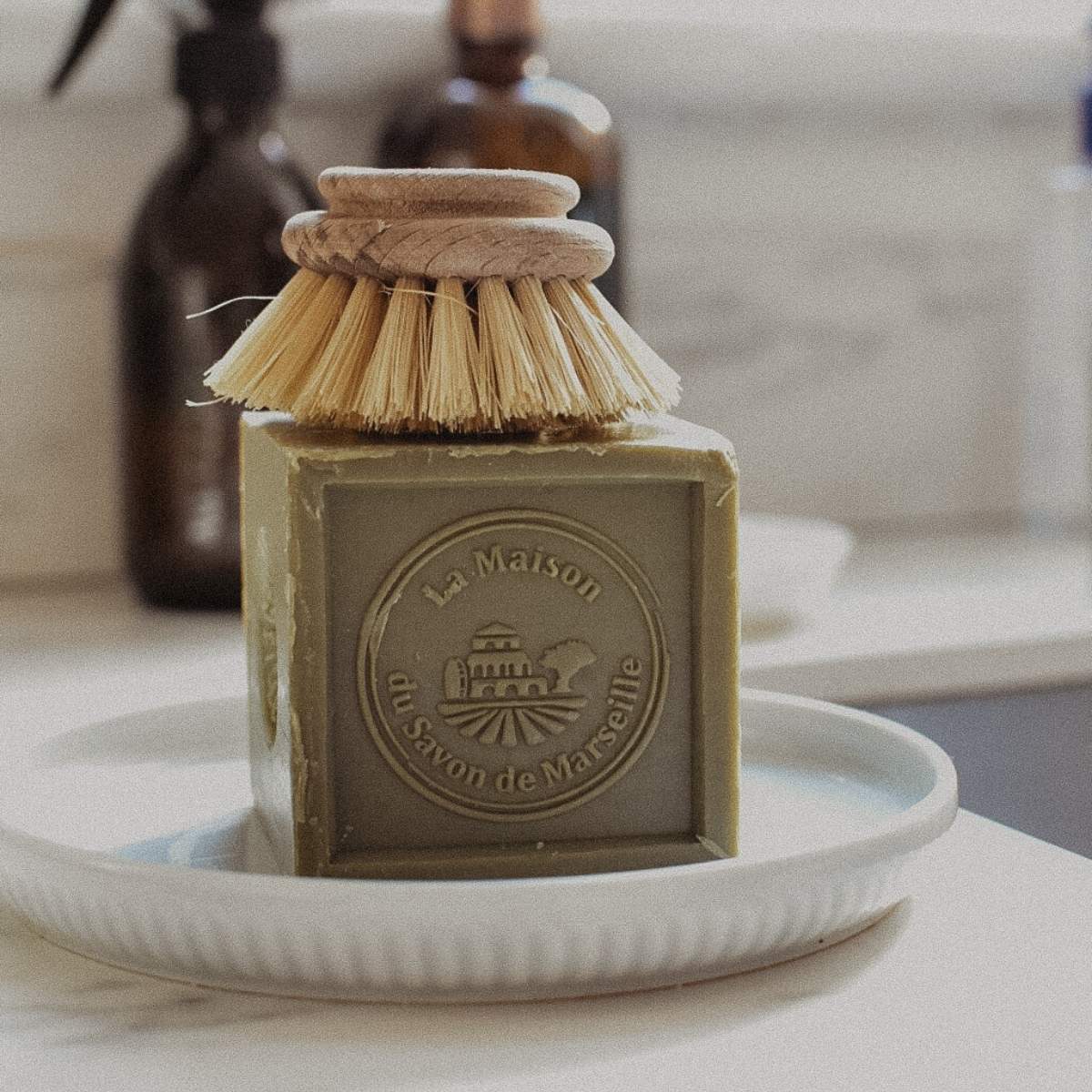 A cube of La Maison du Savon de Marseille soap rests on a ceramic dish, topped with a wooden-handled scrub brush. In the blurred background, brown bottles sit on the white surface.