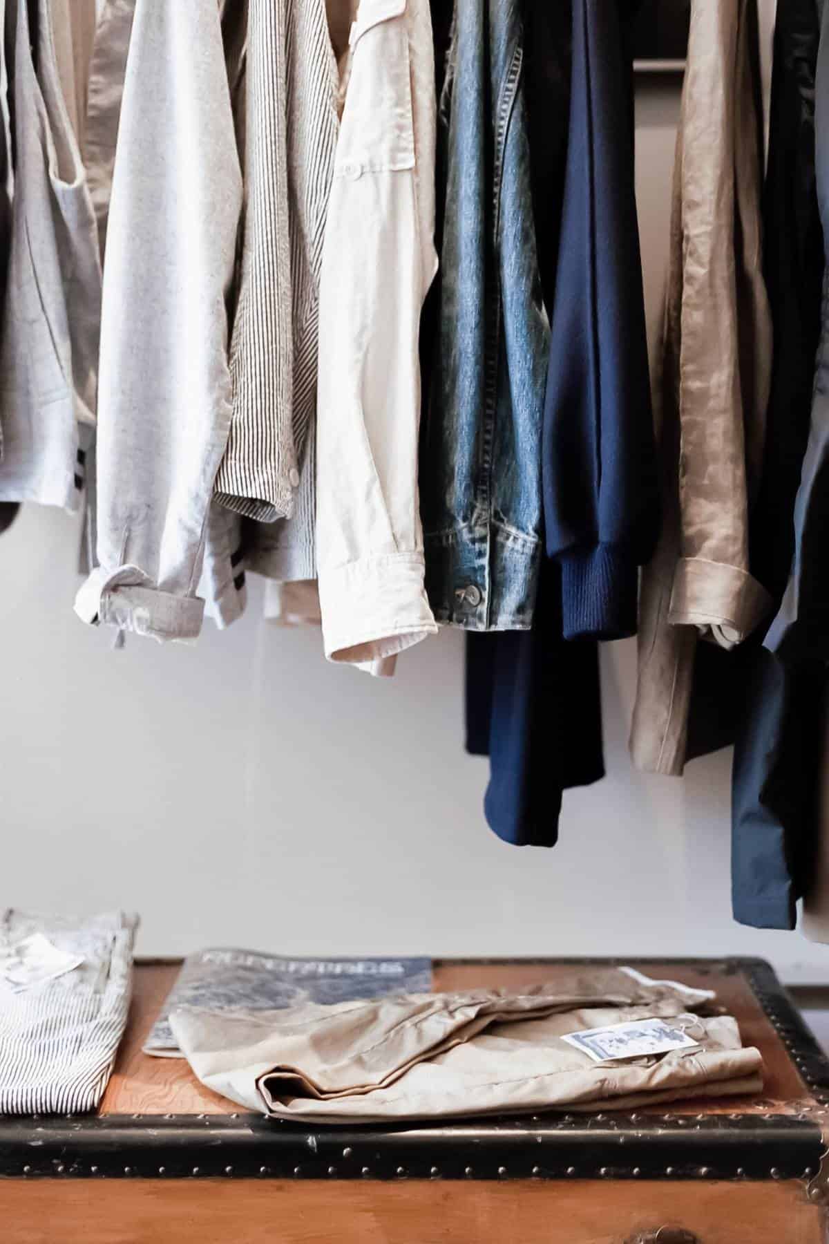 A variety of affordable clothing items hang on a rack, including shirts and a denim jacket. Below them, folded pants and a striped shirt rest on a wooden trunk. The overall color scheme is neutral and denim, making it perfect for building a sustainable wardrobe.