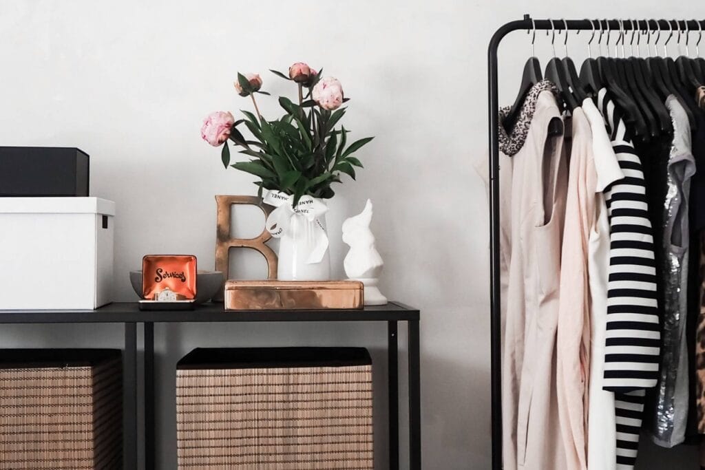 A minimalist room features a black clothing rack showcasing a sustainable and affordable wardrobe. A black shelf holds wicker baskets, a white box, decorative items, and a vase with pink flowers. A small orange box labeled Snacks and a white bust are also on the shelf.