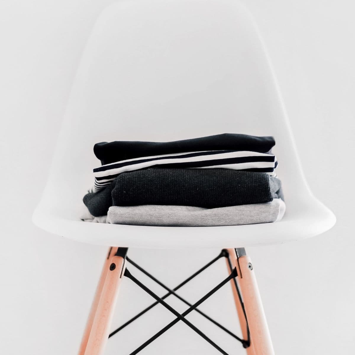 A stack of folded clothes, perfect for a sustainable maternity wardrobe, includes a black, striped, dark gray, and light gray garment. They sit neatly on a modern white chair with wooden legs against a plain white background.