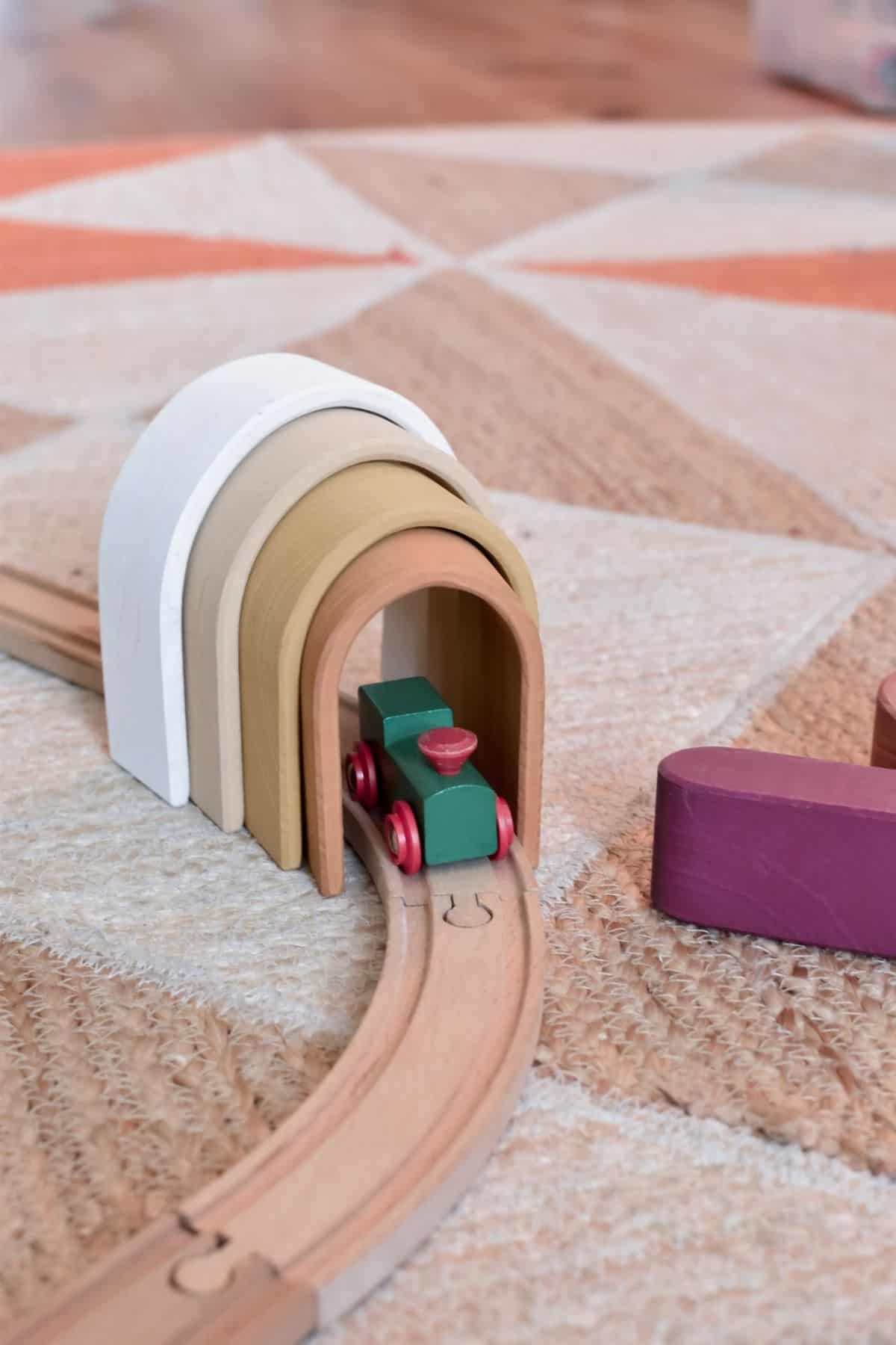 A wooden toy train chugs along a curved track, entering a colorful arch tunnel made by sustainable toy brand Raduga Grez. The arches, stacked in white, beige, mustard, and orange tones.