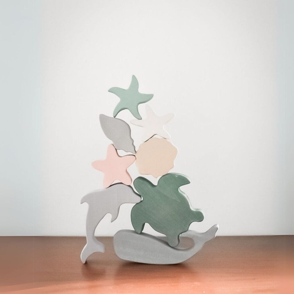 Stacked wooden sea animal shapes made by sustainable toy brand Raduga Grez including a fish, turtle, and starfish in pastel hues, are balanced on a wooden surface. 