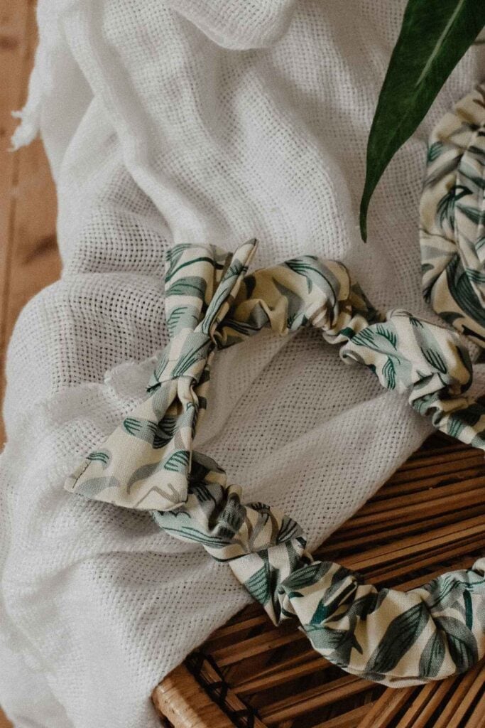 Close-up of a fabric headband with a leaf pattern, perfectly complementing any wardrobe. Resting on a white textured cloth, a green leaf peeks in from the top right corner against a wooden background.