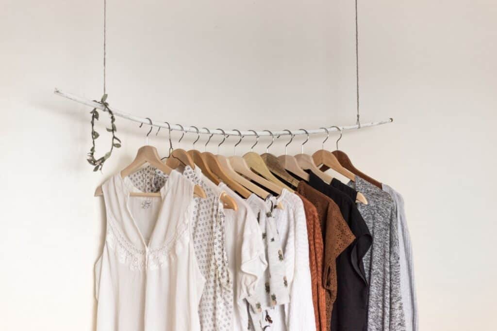 A hanging clothing rack comfortably displays a variety of tops in neutral colors like white, beige, brown, and gray. Suspended by strings with a small plant draping over one end, this setup embodies sustainable style against a plain white wall.