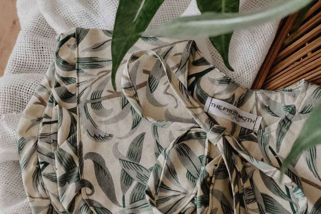A close-up of a fabric with green leaf patterns and a visible tag reading THE FRUMOTH inspires fresh fashion ideas. The fabric is draped on a textured white surface, with a hint of green plant leaf overlapping the image, reminding you to love your wardrobe anew.