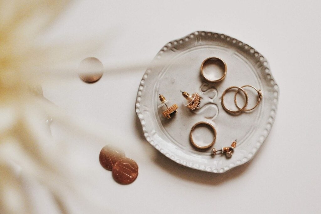 A gray ceramic dish, cherished by mum, holds several pieces of jewelry, including rings and earrings. Two coins rest nearby on the table. The background is softly blurred, adding a warm tone to this sustainably curated scene.