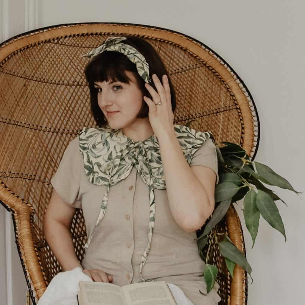 A person with short brown hair sits on a wicker chair, wearing a beige outfit adorned with a large leafy collar and matching headband. They hold a book, contemplating their minimalist wardrobe, while a leafy branch rests against the chair.