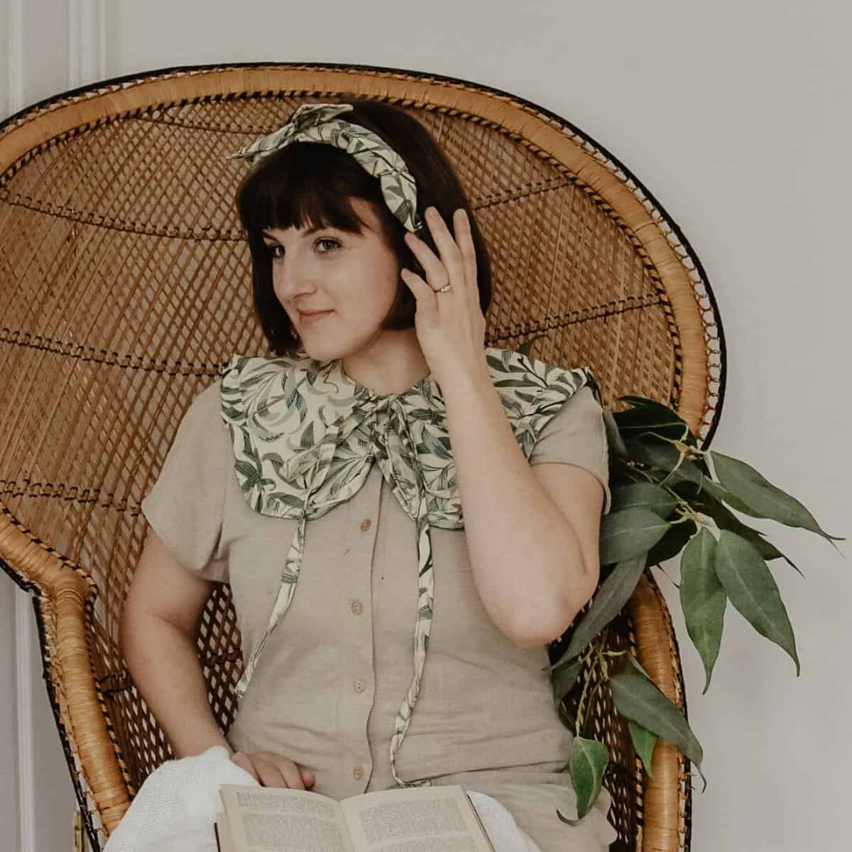 A person with a bob haircut sits in a wicker chair, exuding elegance in a beige outfit adorned with a large leafy collar and matching headband. They hold a book on their lap, with a plant resting on their shoulder—a true testament to how one can love your wardrobe by embracing style and nature effortlessly.