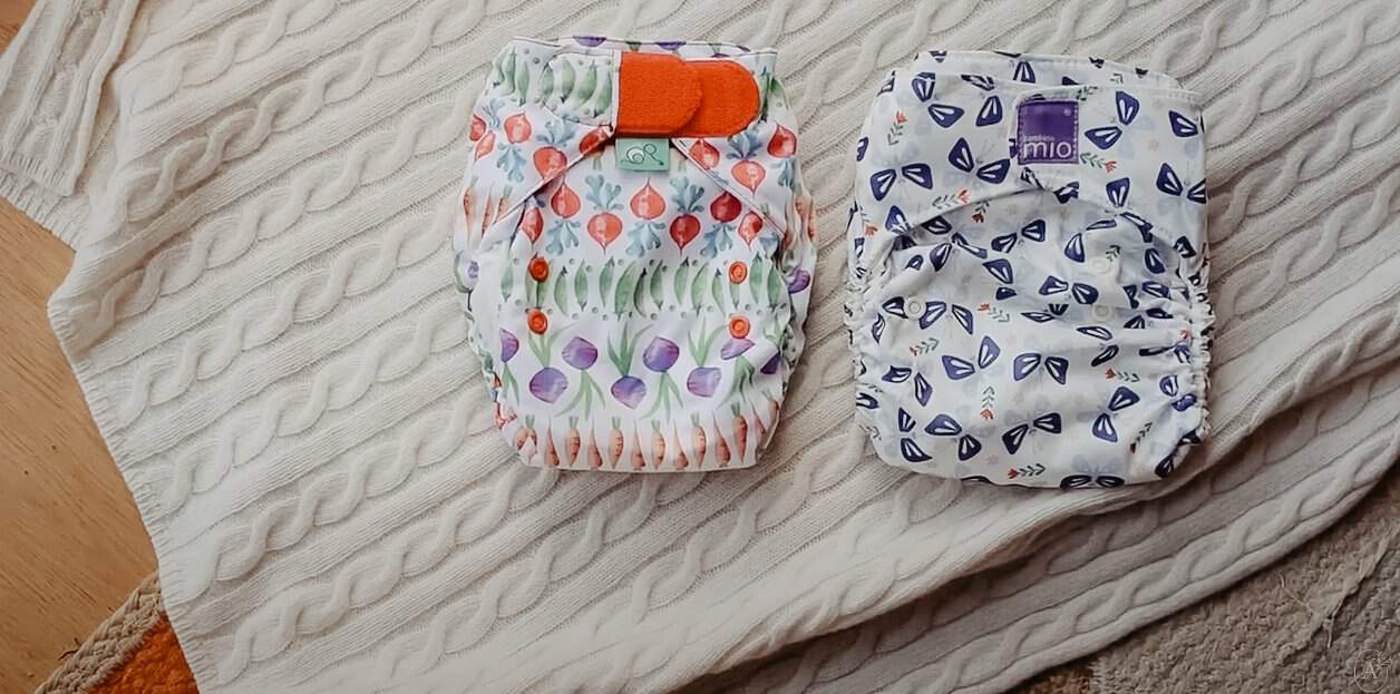 Two colorful cloth nappies rest on a textured beige blanket—one Tots Bots nappy with a vegetable pattern and an orange closure, the other a Bambino Mio nappy showcasing purple and blue butterflies. A wooden floor peeks through, highlighting these delightful cloth nappies.