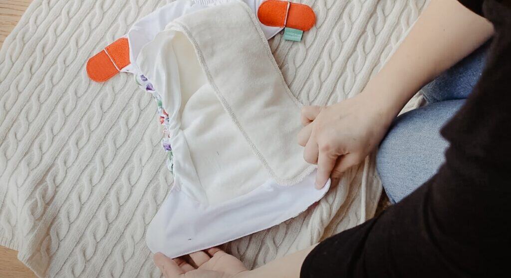 A person prepares a Tots Bots cloth nappy with colorful snaps on a textured wool blanket. Their hands adjust the fabric, showcasing the cloth nappy's interior lining and orange fasteners.