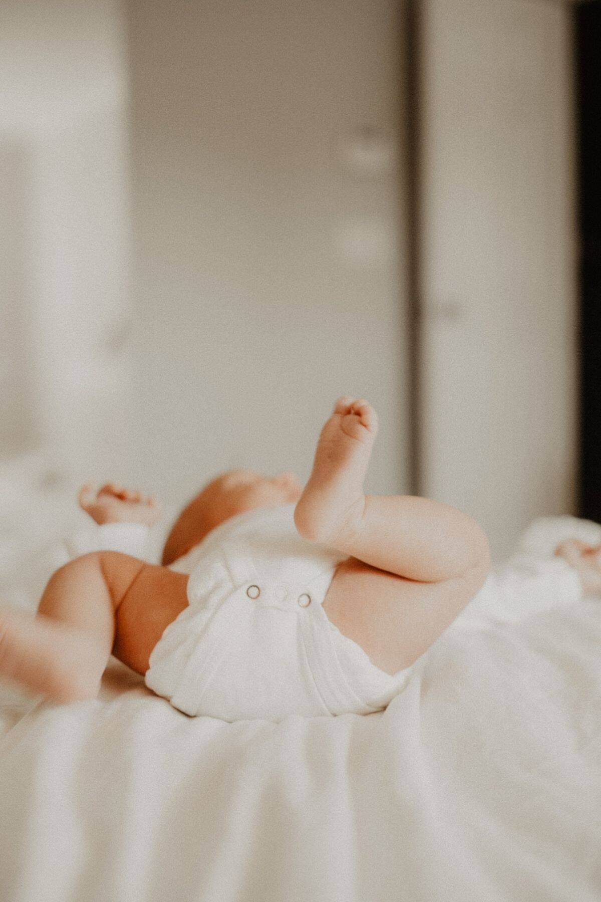 A newborn baby in a white onesie lies on its back on a soft, white bed. The babys legs playfully kick in the air, capturing joy and movement—its a serene snapshot reminiscent of carefree moments parents cherish while traveling with a newborn.