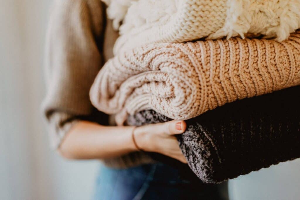 A person holding a stack of folded knitted blankets in beige, light brown, and dark gray, perfect for travelling with a newborn. The cozy sweater they wear complements the softly blurred background, creating a warm, comfortable atmosphere.