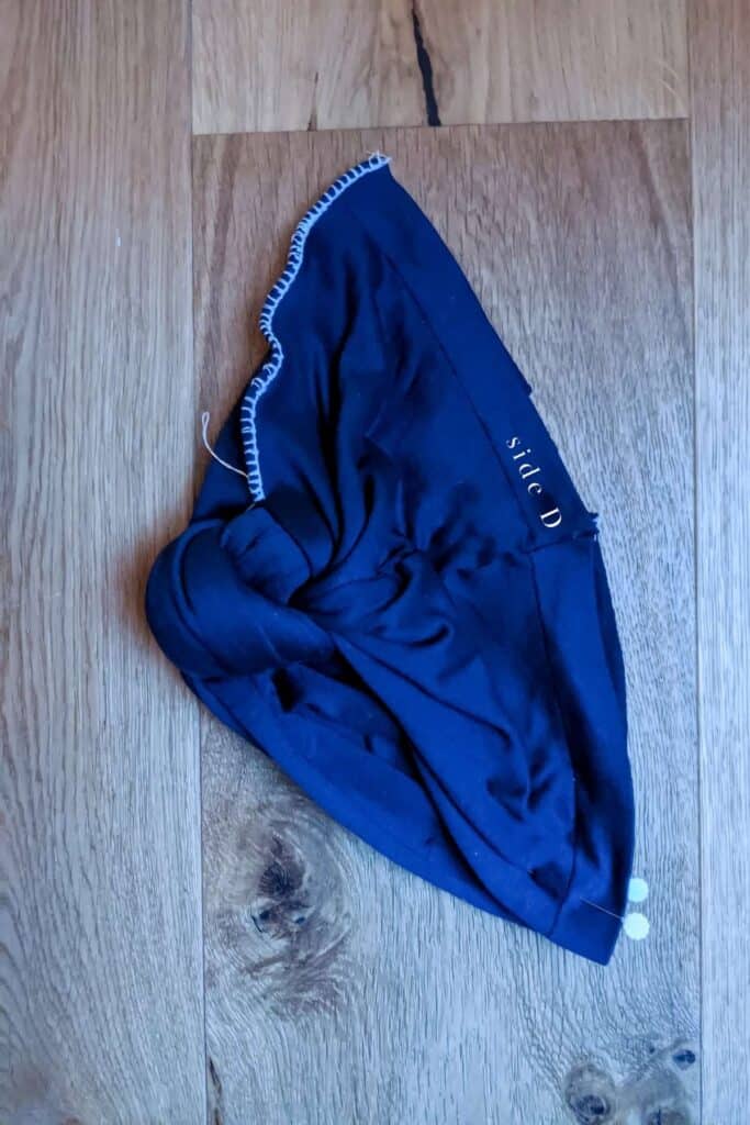 A navy blue, triangle-shaped turban hat with white stitching along the edges lies flat on a wooden surface. One side is labelled side D.