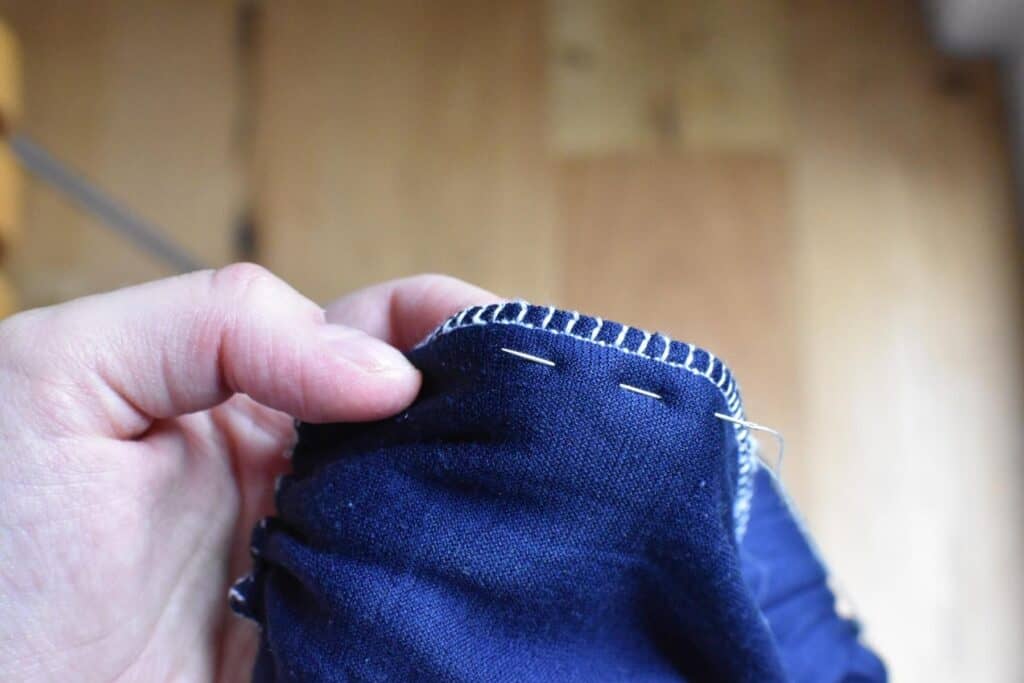 Close-up of a hand sewing the pleats of the turban hat. A needle is threaded with white thread, visible against the fabric. The background offers a blurred view of a wooden floor.