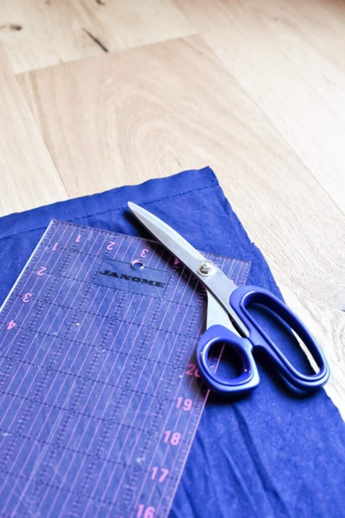 A pair of blue-handled scissors and a transparent measuring ruler rest on a piece of blue fabric, all atop a wooden surface, waiting to transform into a charming baby turban hat.