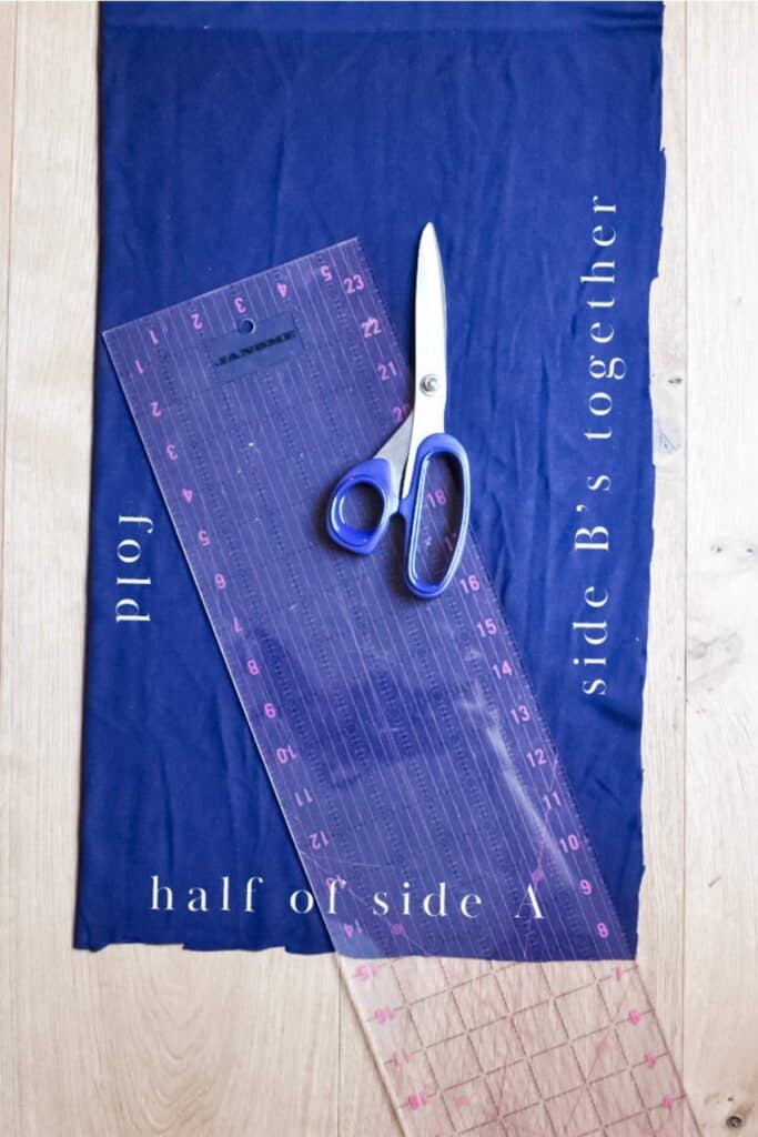 Blue fabric folded with markings for half of side A and side Bs together. A large transparent ruler is placed diagonally, ready to guide your cuts. The setup is on a wooden surface.