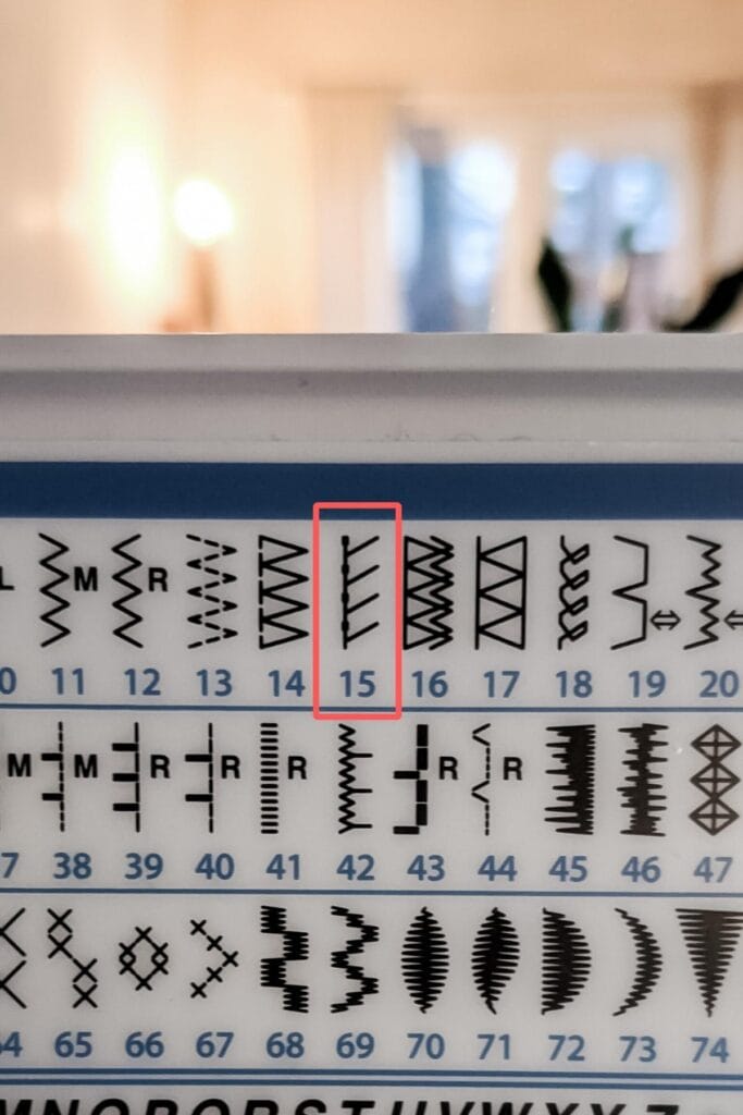 Close-up of a sewing machine stitch pattern chart. Various stitch designs are displayed in rows, each labeled with a number. Pattern number 15, a stretch stitch, is highlighted with a red rectangle. The softly lit room in the background adds warmth to this creative space.