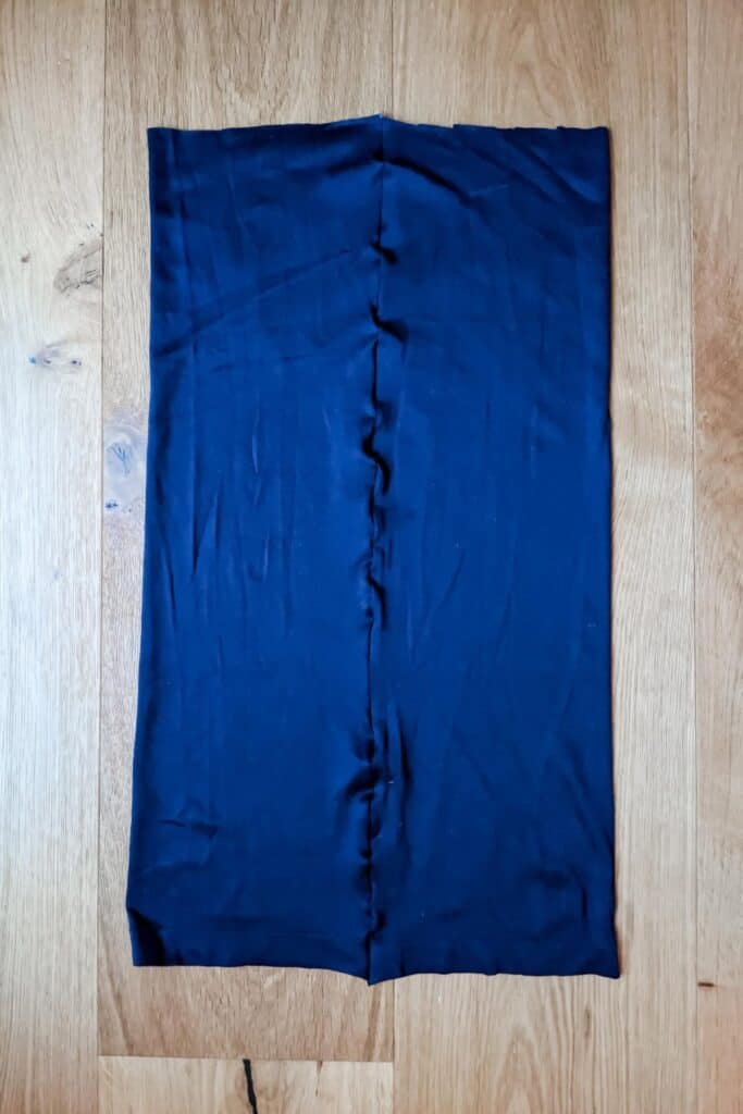 A dark blue fabric tube from an old dress, is laid flat on a wooden floor. The smooth and slightly wrinkled material features a visible seam down the center.