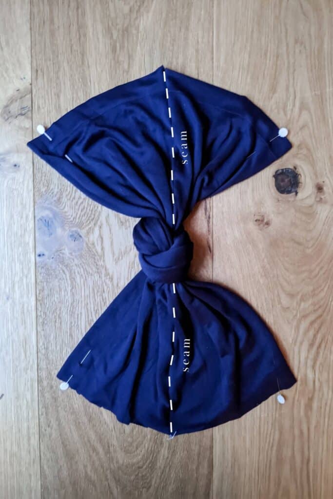 The baby turban hat resembles a bow tie, showcasing stitched lines and the word seam labeled down the centre, with small white pins at the corners. 