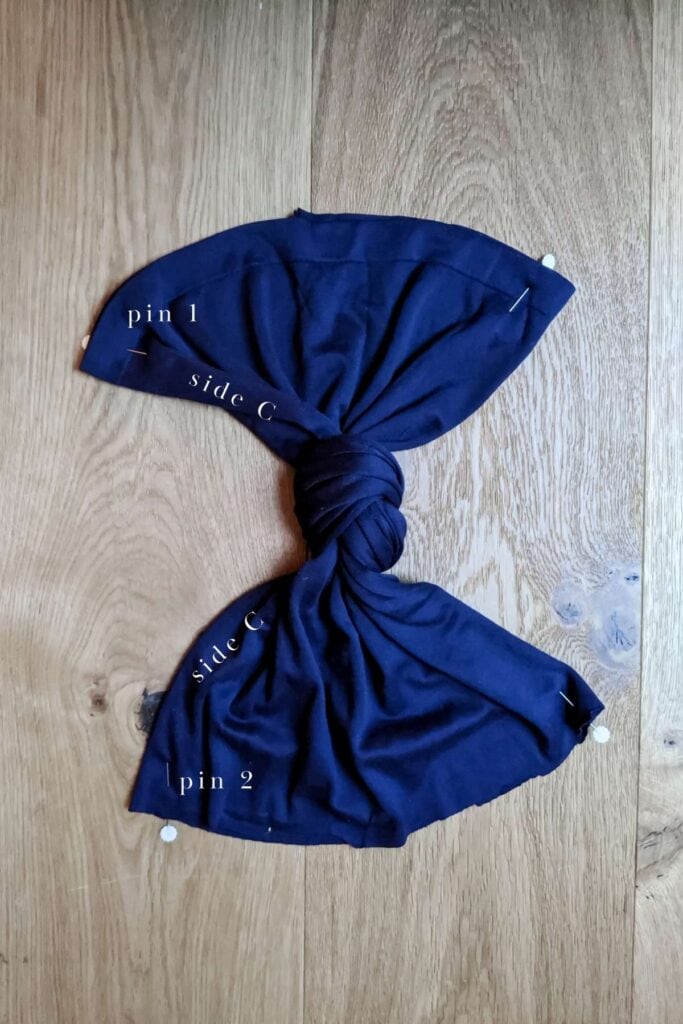 A dark navy piece of fabric, perhaps an old dress, is laid on a wooden surface, knotted in the center like a DIY project for a baby turban hat. The fabric features labels like pin 1, side C, and pin 2 marked on its sections.