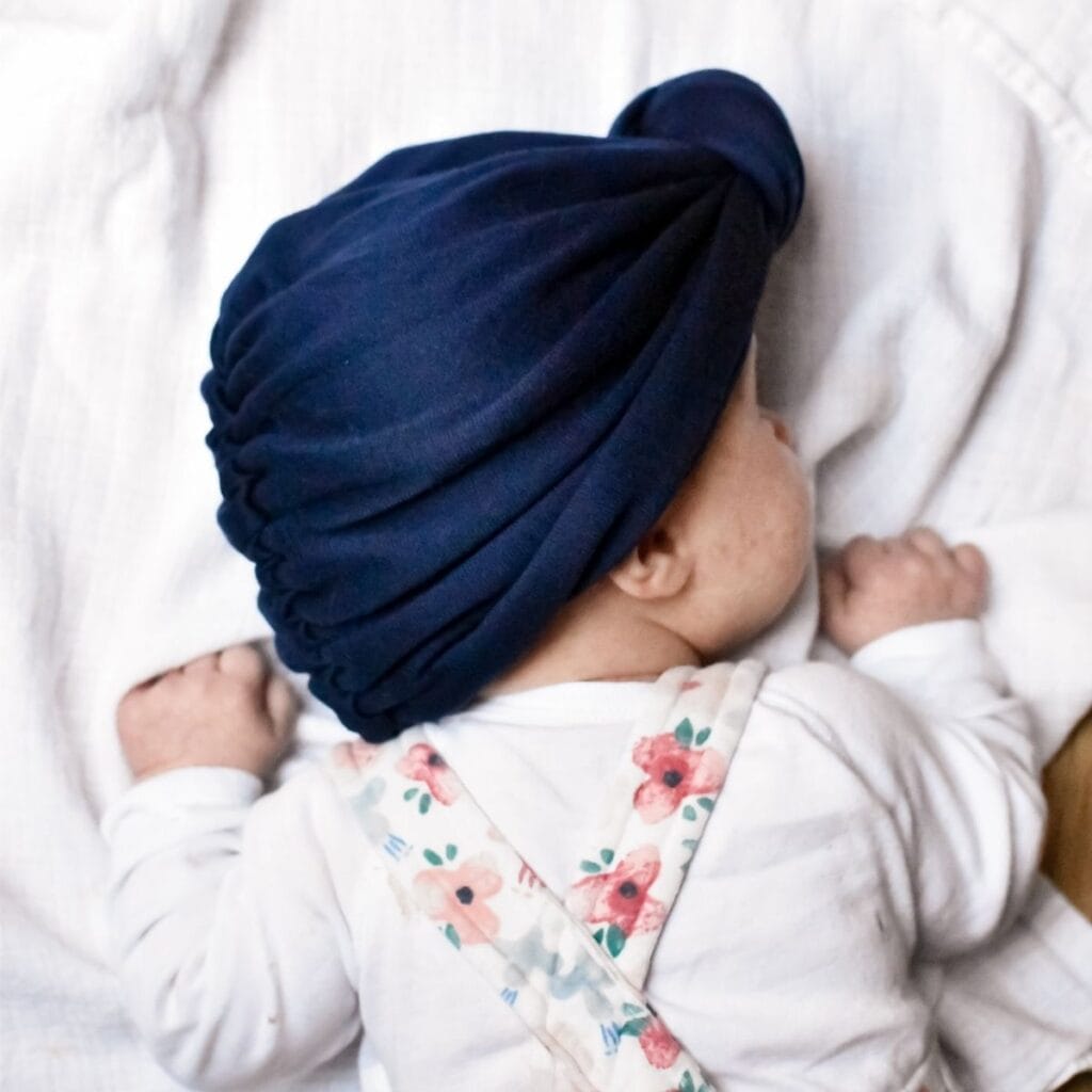 A baby wearing a white outfit with floral patterns and a navy-blue turban made from an old dress lies on a white blanket, facing away. The baby has both arms bent and rests on their stomach.