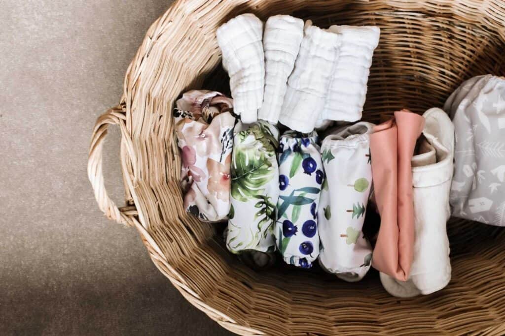A wicker basket filled with neatly arranged cloth nappies showcases various colorful patterns, including floral, blueberries, and abstract designs. This charming display serves as a guide to the types available, all on a neutral-colored surface.