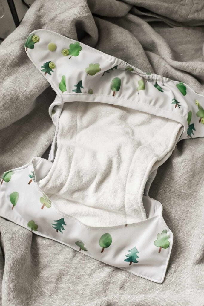 A cloth nappy with a whimsical tree pattern is open and laid out on a gray fabric surface. This charming design features illustrations of various green trees and plants, complemented by a white absorbent lining inside.