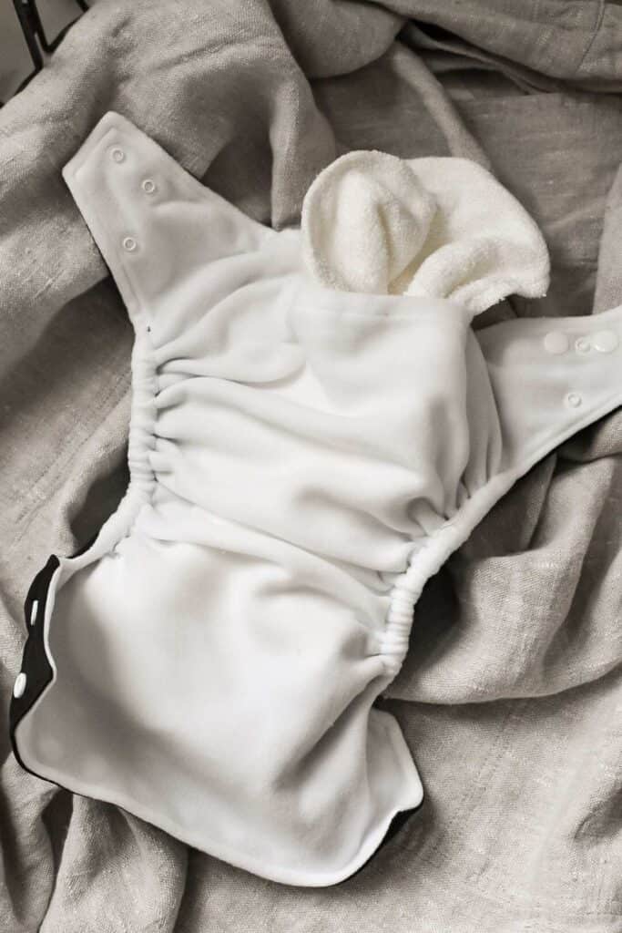 A white cloth nappy with snap fasteners lies open on a textured gray fabric. The nappy appears soft and reusable, featuring elasticized edges and a partially visible absorbent insert.