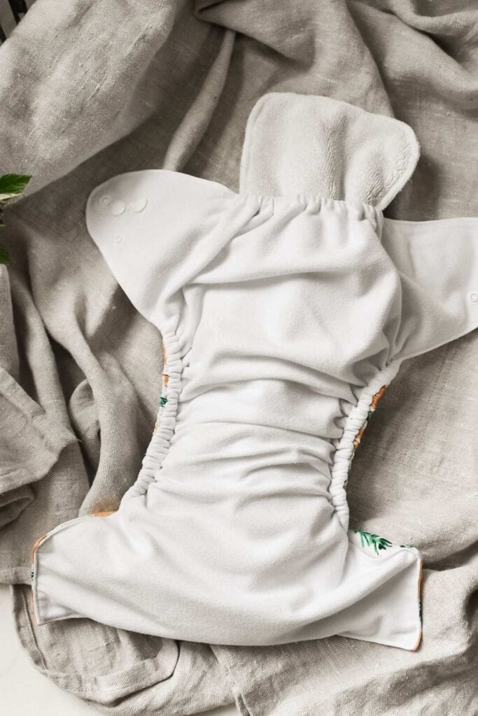 This cloth nappy, featuring a soft white fabric set against a textured light gray background, showcases elastic gatherings and a flap with snap buttons. The edges boast green and orange accents, beautifully illustrating the types of cloth nappies available in our complete guide.