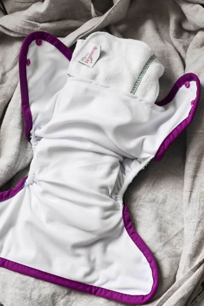 A cloth nappy is open and laid out on a textured fabric surface. The diaper, part of various types of cloth nappies, is predominantly white with purple trim and features a soft material interior. Snap buttons are visible for closure.
