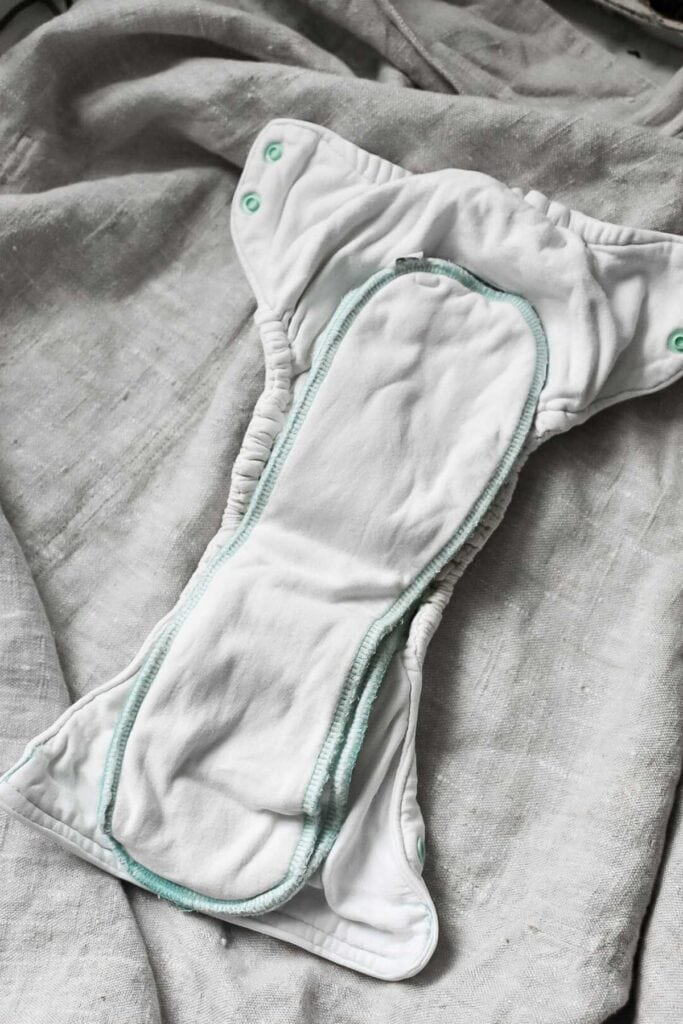 A reusable cloth nappy with a white absorbent insert is laid out on a textured grey fabric surface. The nappy features green snaps and elastic sides, exemplifying one of the advanced types of cloth nappy designs.