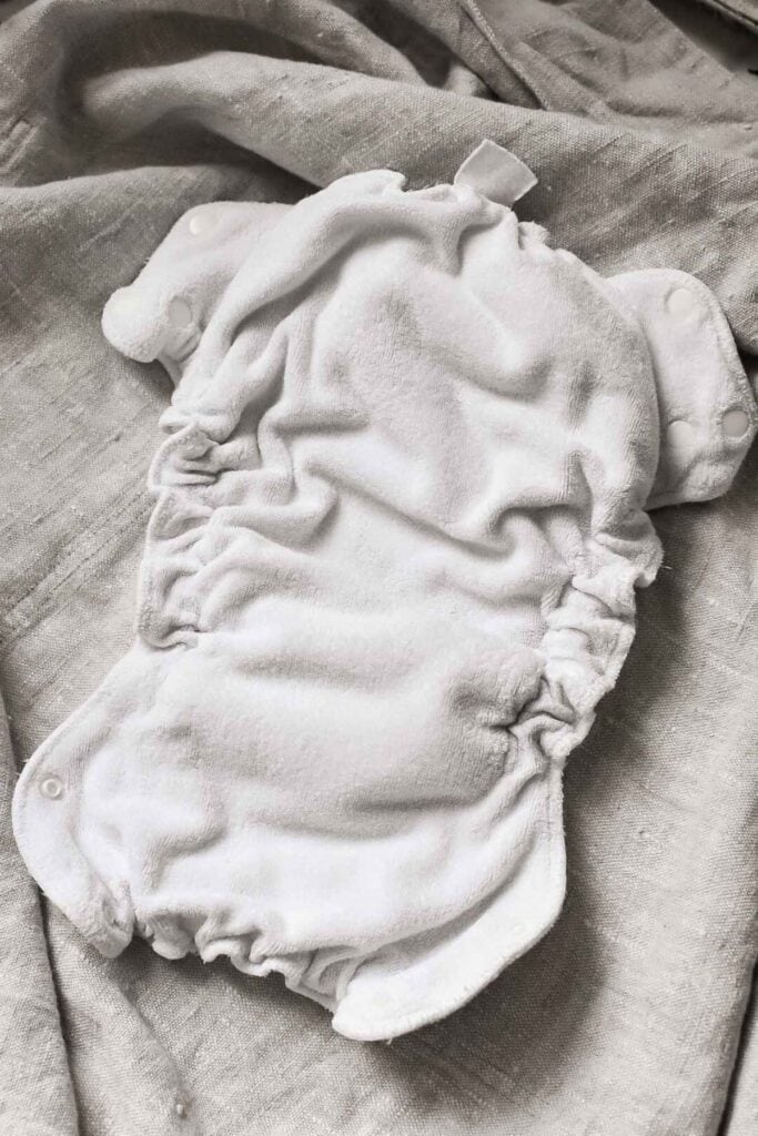A soft, white cloth nappy with elastic sides and snap buttons lies open on a textured gray fabric background, exemplifying one of the types featured in our complete guide.