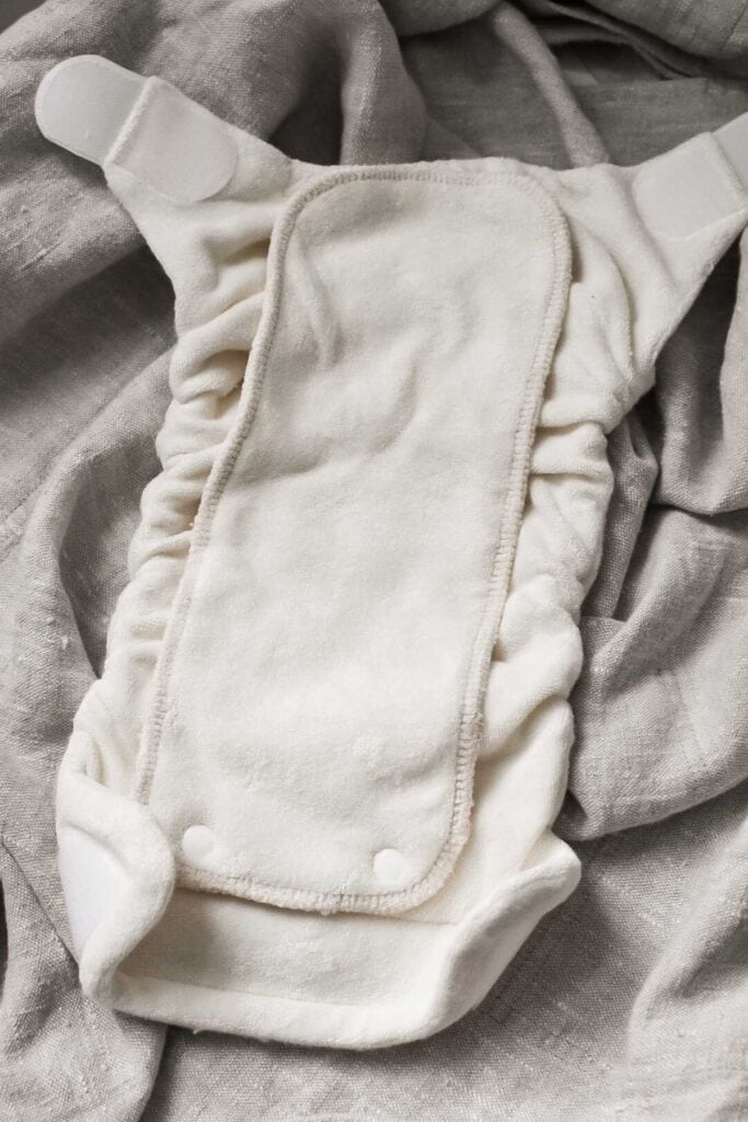 A beige cloth nappy with Velcro straps is laid out on a textured gray fabric background. This type of cloth nappy features elastic sides and snap buttons at the bottom, showcasing a complete guide to convenience and comfort.