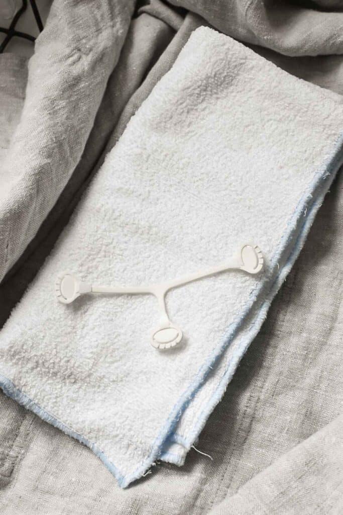 A folded white towel rests on light gray fabric, resembling the soft folds of a cloth nappy. A small, white plastic massage tool with three prongs is placed on top of the towel, completing this soothing setup.