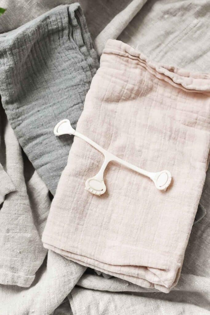 A white plastic facial lymphatic drainage tool rests on soft, folded gray and beige cloths, creating a soothing and minimalist aesthetic reminiscent of a gentle guide to cloth nappy care.