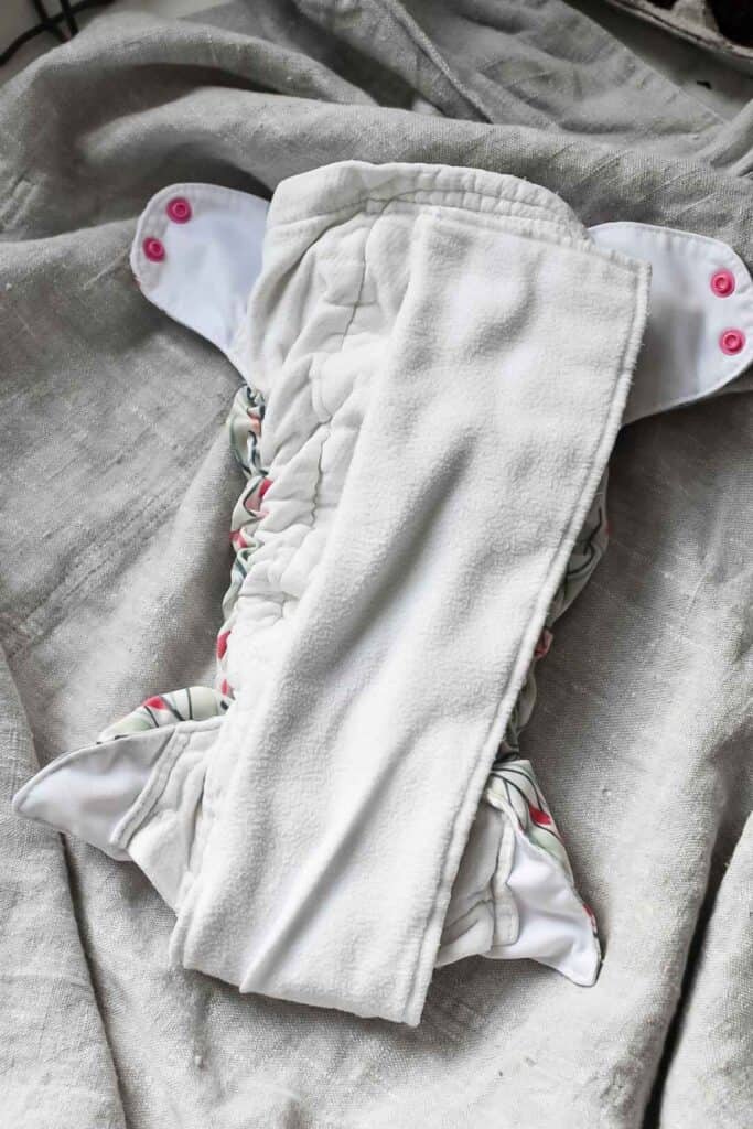 This cloth nappy features a soft white insert and vibrant floral-patterned edges against a gray surface. It has pink button fasteners at the sides, offering a delightful style in the world of nappy types.