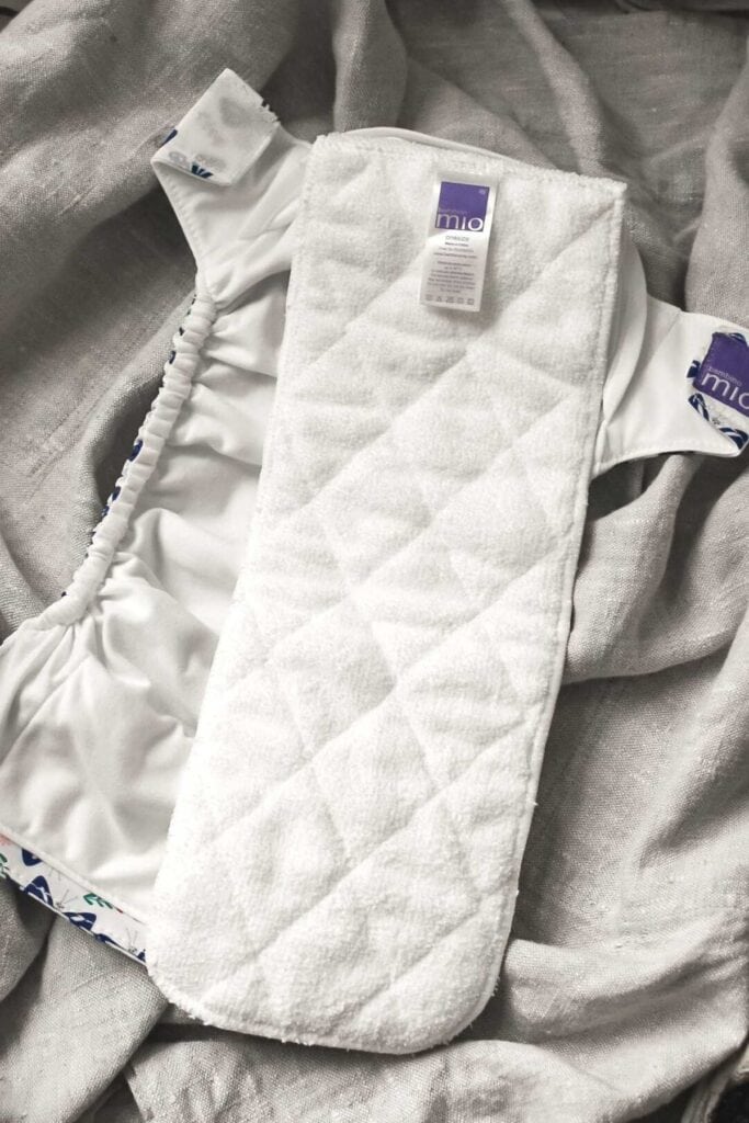 Discover the versatility of our cloth nappy, featuring a white quilted insert nestled on a gray fabric backdrop. This diaper, complete with elastic edges and vibrant patterned tabs, serves as a perfect guide for parents exploring different types of reusable options.