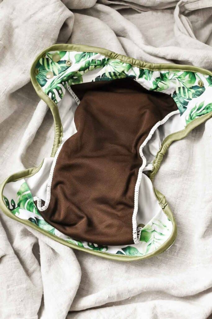 A reusable cloth nappy features a brown inner lining and a green leafy pattern on the edges and outer fabric. Perfectly captured on a soft, light gray fabric background, this design exemplifies the elegance found in various types of cloth nappies.